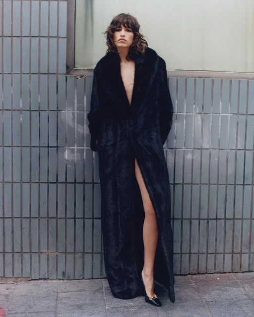 Adult woman posing in a long black fur coat with a thigh-high slit, wearing black heels against a gray tiled wall outdoors.