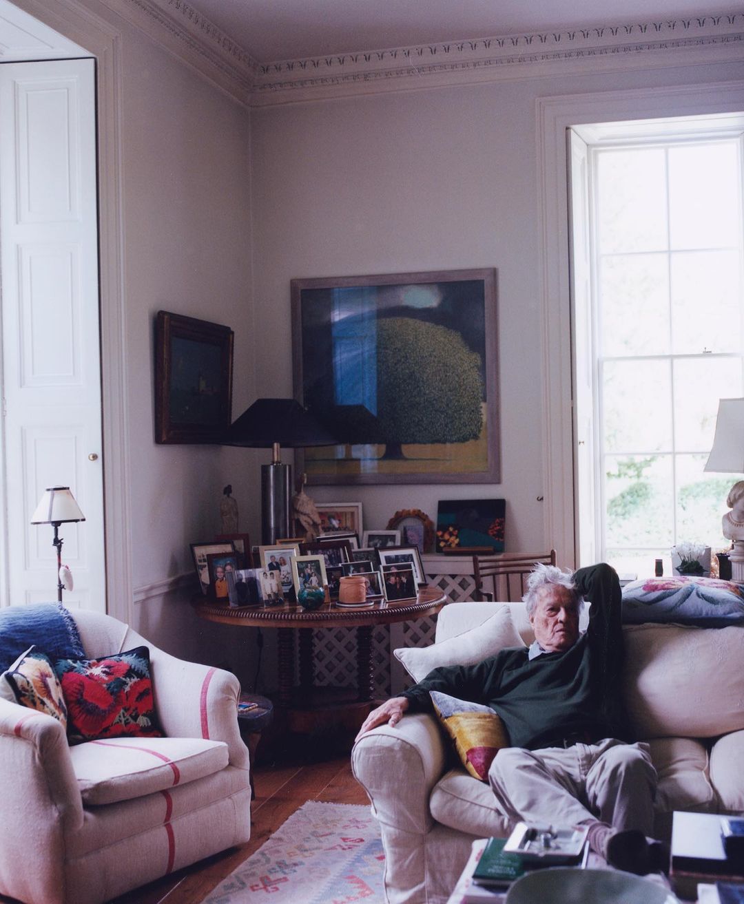 An elderly man seated on a white sofa in a brightly lit traditional living room, photographed by Charlie Gates.