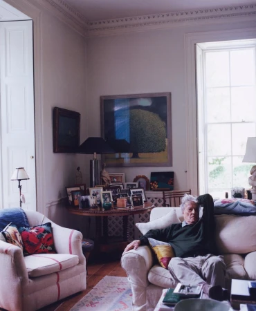 An elderly man seated on a white sofa in a brightly lit traditional living room, photographed by Charlie Gates.