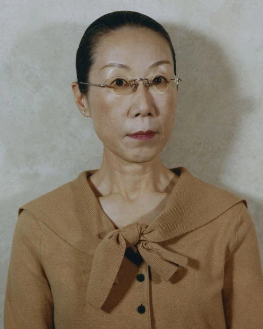 Portrait of an Asian woman wearing spectacles and a tan cardigan with a bow, set against a neutral textured backdrop in a studio.