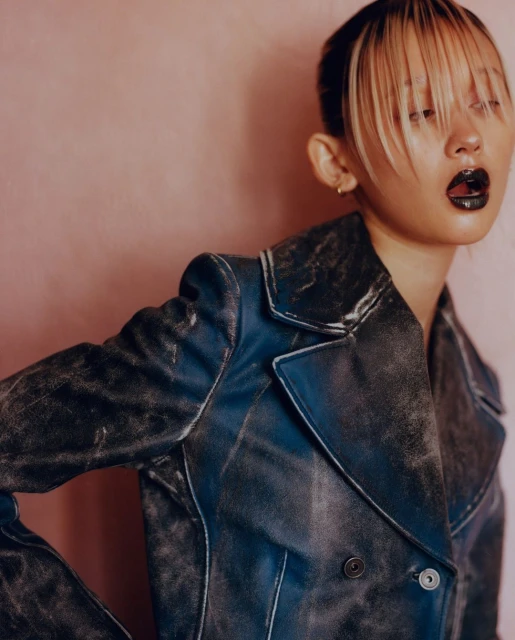 Close-up portrait of a young adult woman with dyed blonde hair, wearing a worn blue leather jacket against a light pink background.