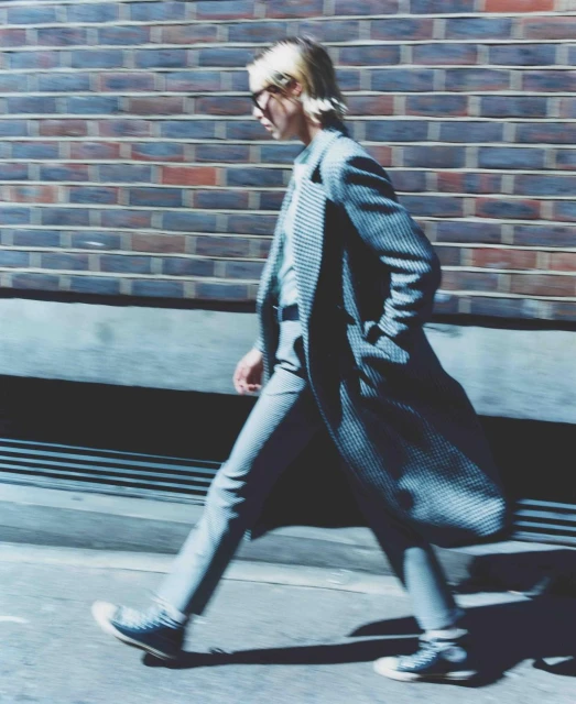 Fashionable adult male in a striped suit and sneakers walks along a city sidewalk past a brick wall in full sunlight.