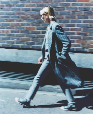 Fashionable adult male in a striped suit and sneakers walks along a city sidewalk past a brick wall in full sunlight.