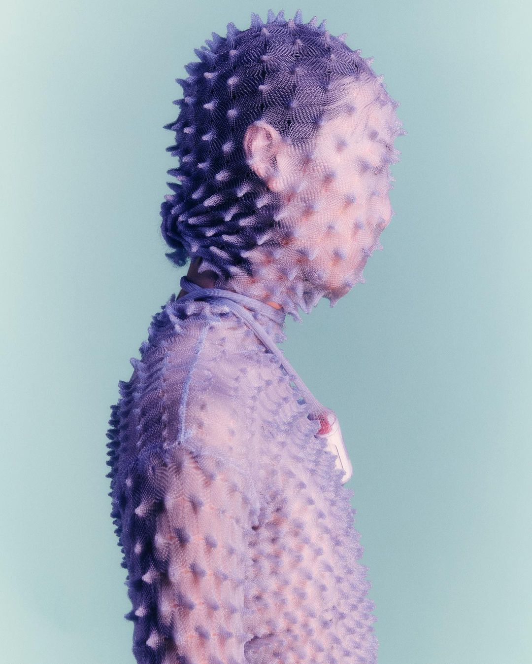 Person in spiky purple fashion, wearing head-to-toe outfit with textured spikes, against a soft blue backdrop, side profile.