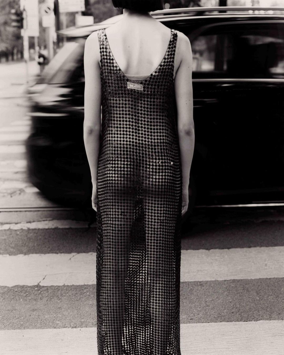 Adult female in maxi dress standing in a crosswalk in a monochrome fashion photograph with a vehicle passing by in the background.