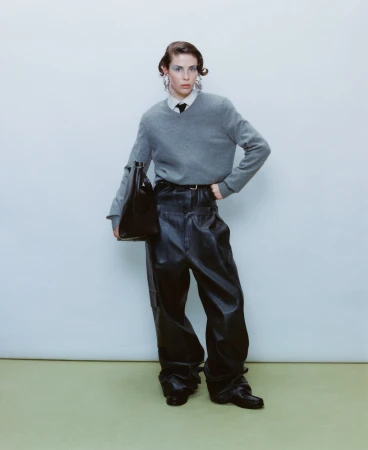 Model in grey sweater and black leather trousers holding a handbag against a plain background