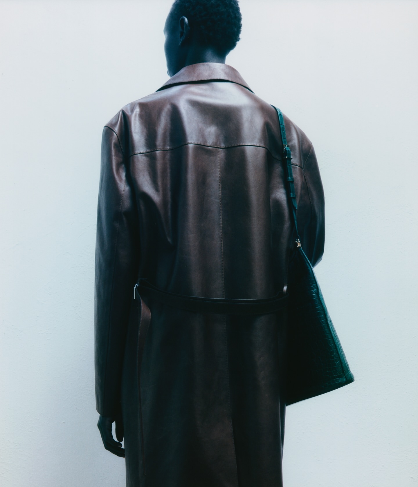 Back view of a Black male in a brown leather trench coat with a dark bag over his shoulder, posing against a light neutral background.