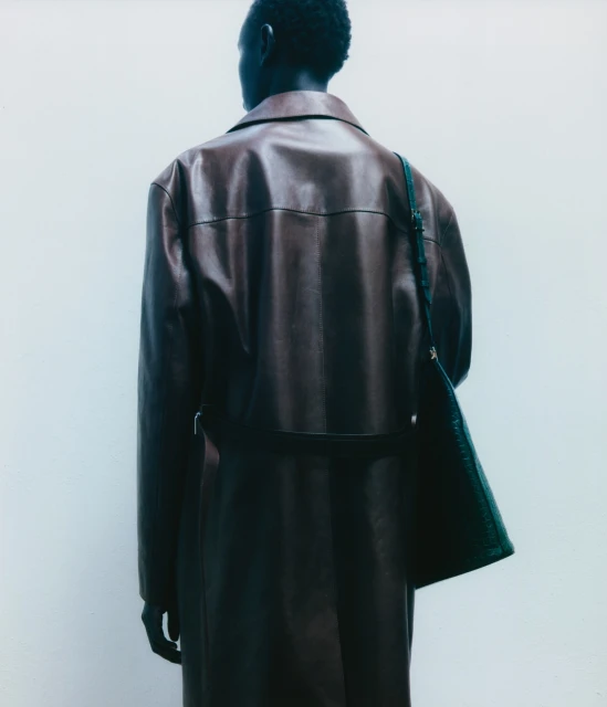Back view of a Black male in a brown leather trench coat with a dark bag over his shoulder, posing against a light neutral background.