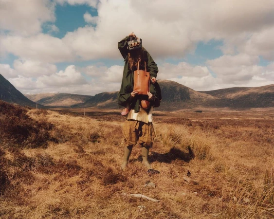 Woman gives a child a piggyback ride through a grassy field with rolling hills and a cloudy sky in the background.