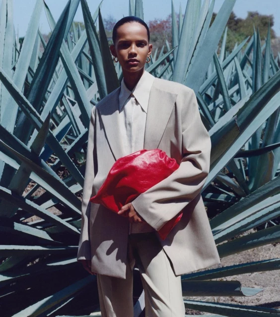 Fashion shot of a woman in a beige suit holding a red bag in a natural setting, featuring large agave plants in the background.