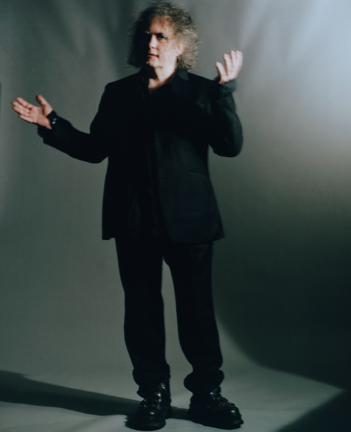 Full body portrait of an adult male with curly hair wearing a black suit and boots against a grey background in a photo studio.