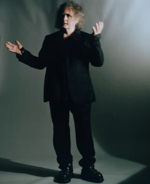 Full body portrait of an adult male with curly hair wearing a black suit and boots against a grey background in a photo studio.
