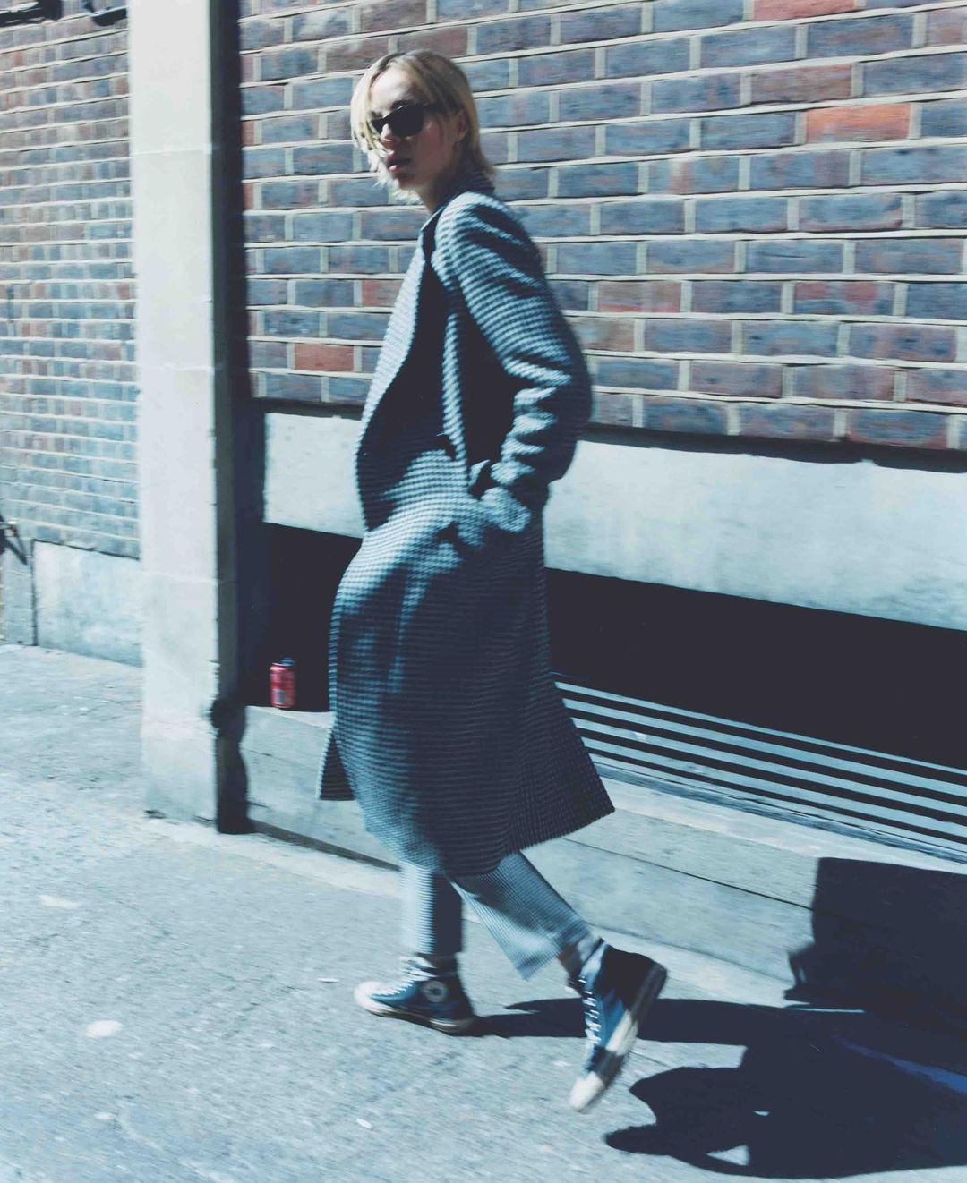 Fashionable adult woman walking in an urban setting. She wears a long coat, light pants, and sneakers against a brick wall backdrop.