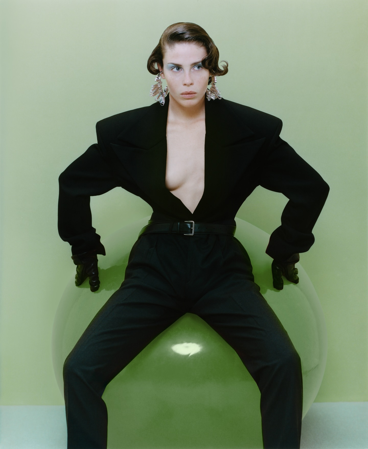 A stylish woman in a black suit strikes a pose, seated on a large green balloon against a light green backdrop in a studio.