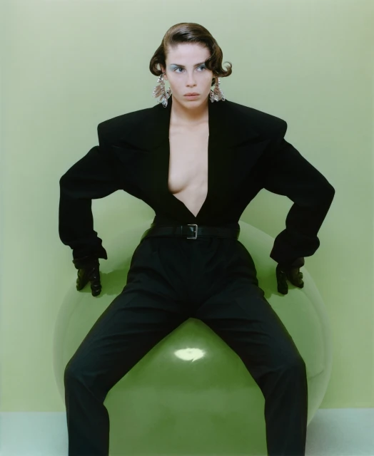 A stylish woman in a black suit strikes a pose, seated on a large green balloon against a light green backdrop in a studio.