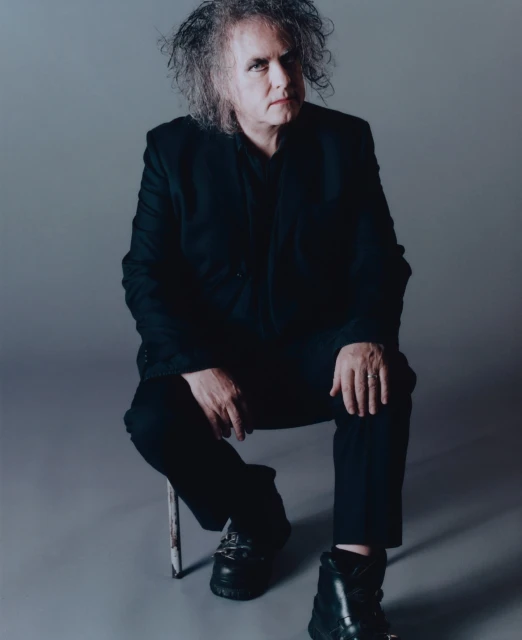 Adult male seated on a stool in a photo studio. He has curly hair and is wearing a black suit and boots against a neutral gray backdrop.
