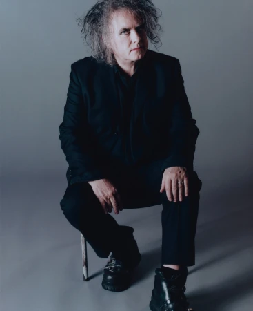 Adult male seated on a stool in a photo studio. He has curly hair and is wearing a black suit and boots against a neutral gray backdrop.