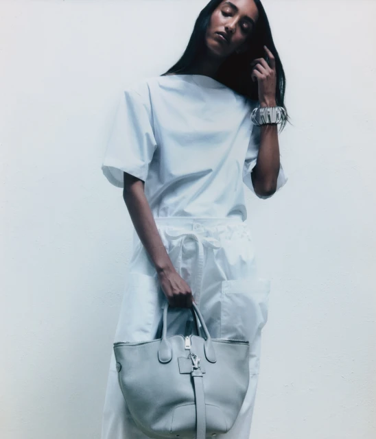 Woman in white outfit holding grey tote bag against a white background. Silver bracelet accentuates the minimalist fashion style.