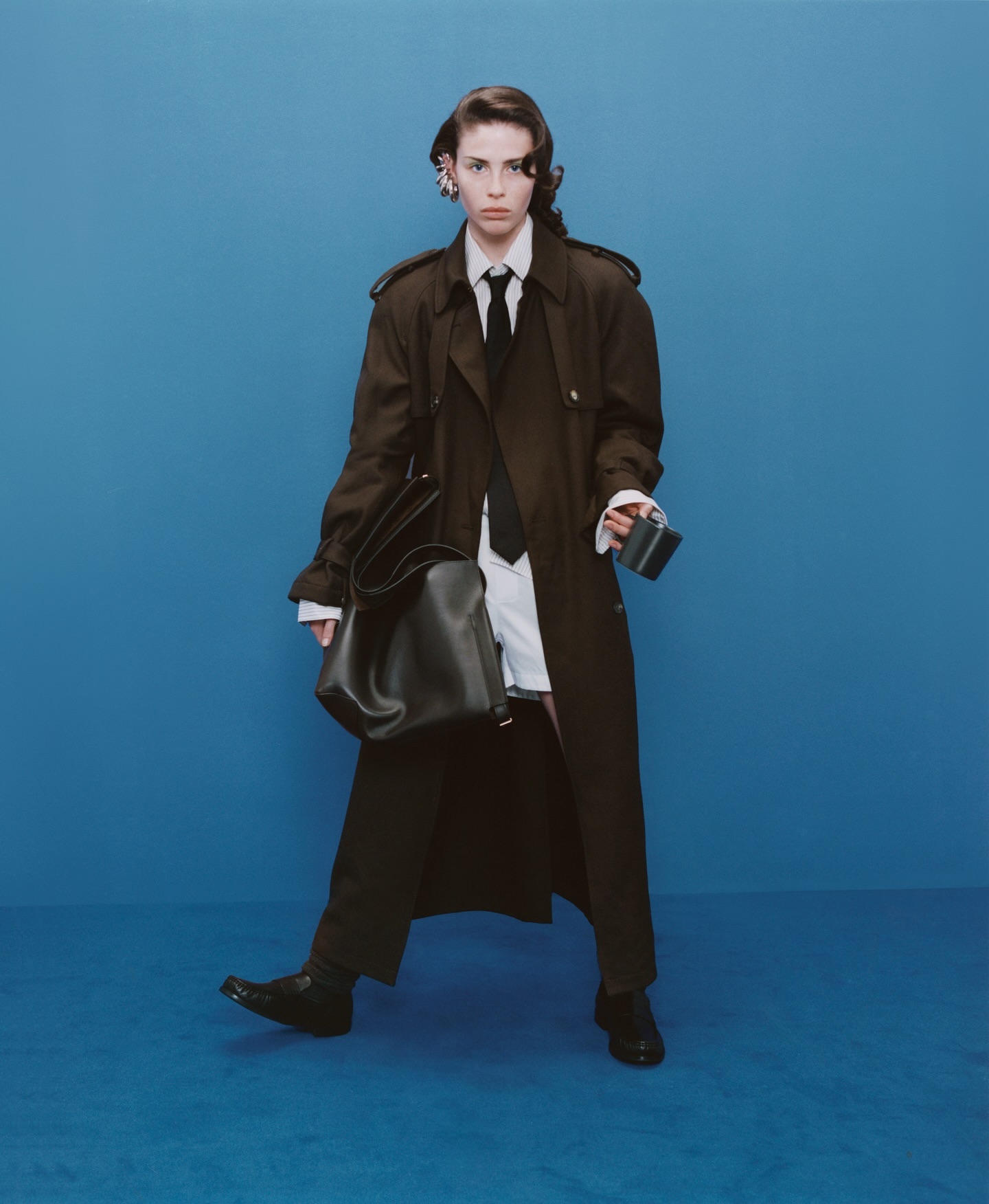 A woman poses against a blue backdrop wearing a brown trench coat, white shirt, black tie, black pants, and black shoes.