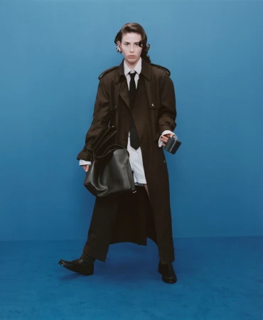 A woman poses against a blue backdrop wearing a brown trench coat, white shirt, black tie, black pants, and black shoes.