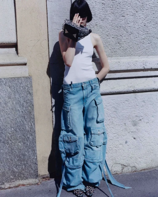 A slender model with a bob poses in baggy denim cargo pants and a white tank top, accented by a small handbag and patterned shoes.