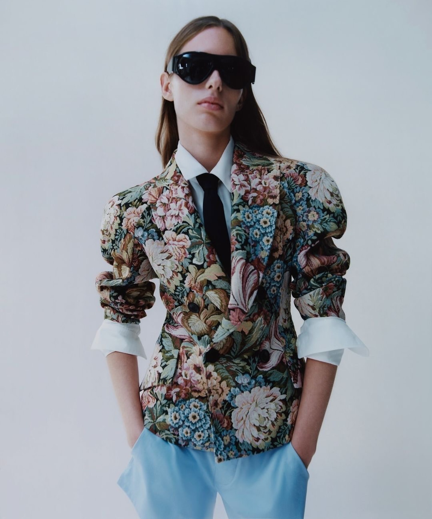 Model in a floral blazer, light blue trousers, sunglasses and a tie, posing in a studio setting against a white backdrop in a fashion editorial.