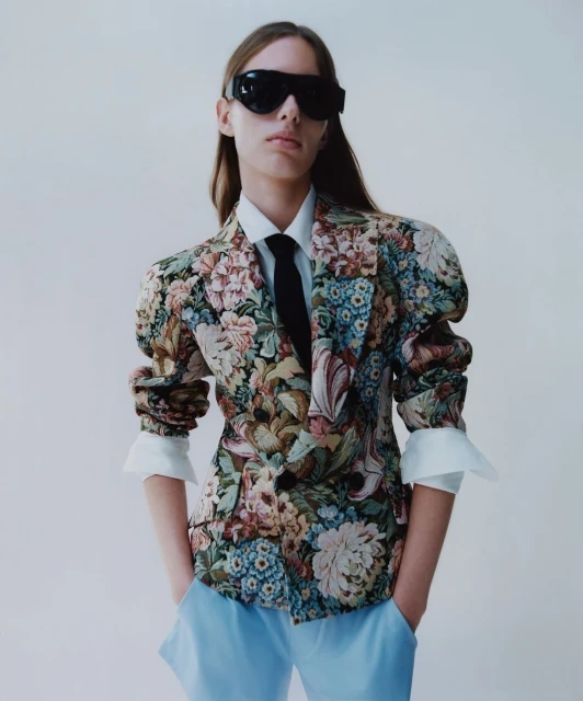 Model in a floral blazer, light blue trousers, sunglasses and a tie, posing in a studio setting against a white backdrop in a fashion editorial.