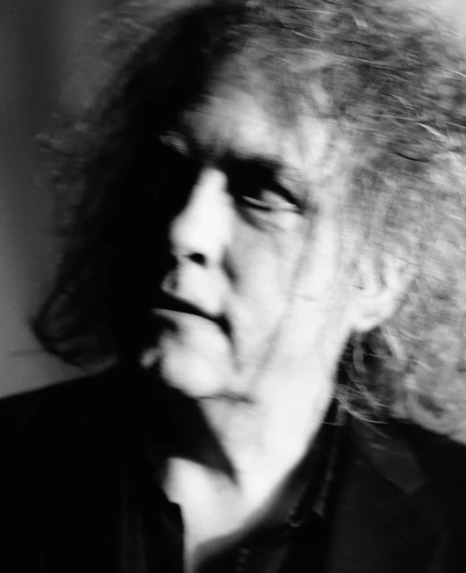 Monochrome portrait of an adult male with curly hair, dark suit, and dramatic lighting creating deep shadows and a blurred effect.