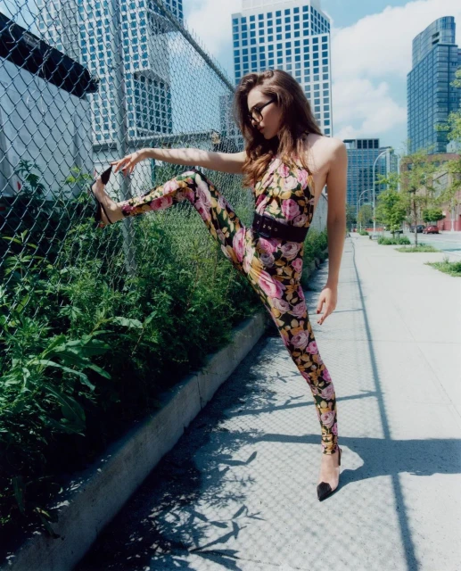 A woman in a floral jumpsuit poses with one leg up against a chain-link fence, wearing heels, with city buildings in the background.