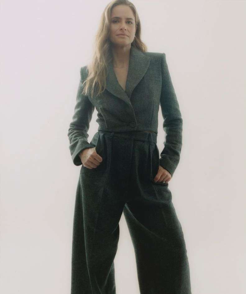 A full-length studio portrait of a woman with long hair, wearing a grey jacket and wide-leg pants, standing with her hands in her pockets.