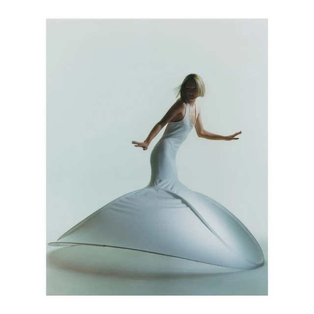 Woman in a form-fitting white gown with a dramatically flared skirt strikes an elegant pose in a bright studio setting.