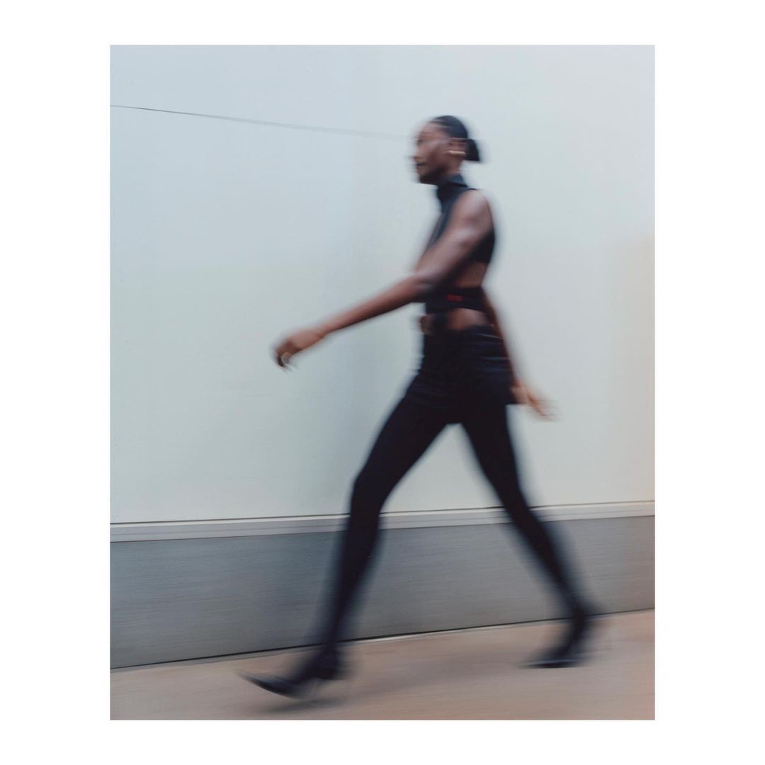 A blurry side view of an adult woman walking against a neutral wall. The subject is wearing a black, cut-out top and matching pants.