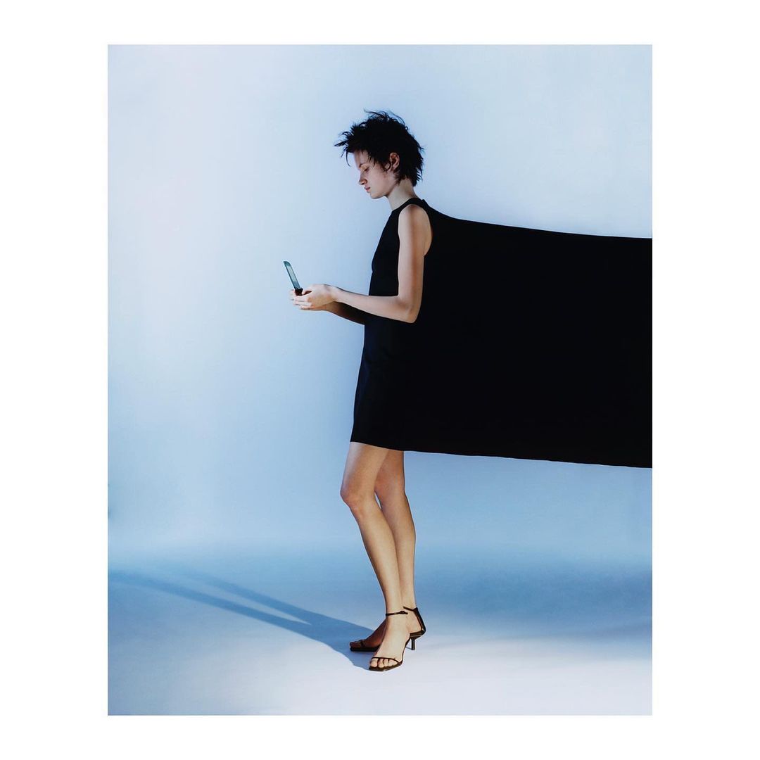Woman in a black dress using her phone in a studio with bright lighting, casting a shadow. The dress has a dramatic extension.
