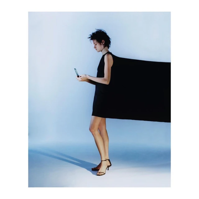 Woman in a black dress using her phone in a studio with bright lighting, casting a shadow. The dress has a dramatic extension.