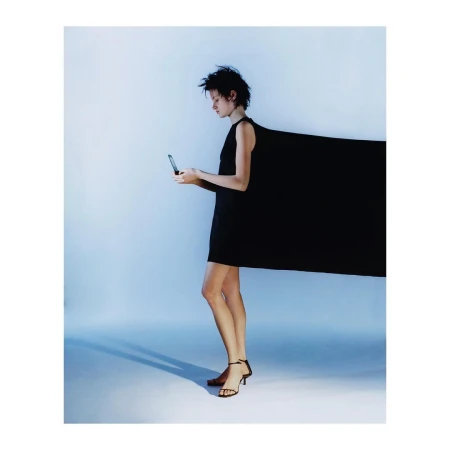 Woman in a black dress using her phone in a studio with bright lighting, casting a shadow. The dress has a dramatic extension.