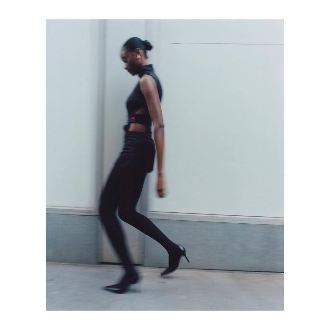 Adult female model in black top and leggings walking outdoors. Fashion shot with motion blur, set against a neutral urban background.