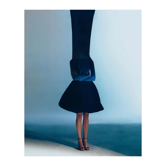 A woman with crossed arms stands in a black dress and strappy sandals against a gradient blue-gray background, creating an elegant fashion image.
