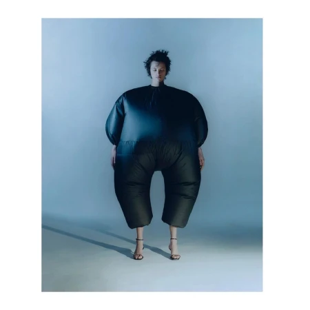 A person wearing a dark, oversized puffy jumpsuit and thin strap heels standing against a neutral studio backdrop in London.