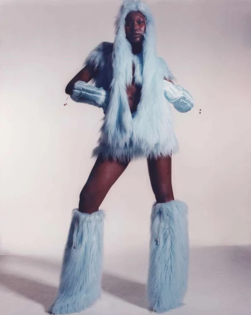 Dark-skinned woman in light blue faux fur outfit, including hood, vest, gloves, mini skirt, and leg warmers against a white backdrop.