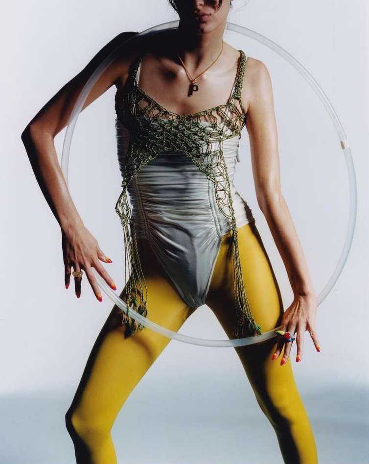Model poses in a light grey one-piece swimsuit, yellow tights, and a green crocheted top while holding a transparent hula hoop.