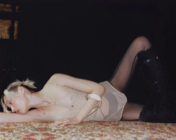 Model lying on patterned carpet wearing nude bodysuit, black tights, and fluffy dark boots against dark background