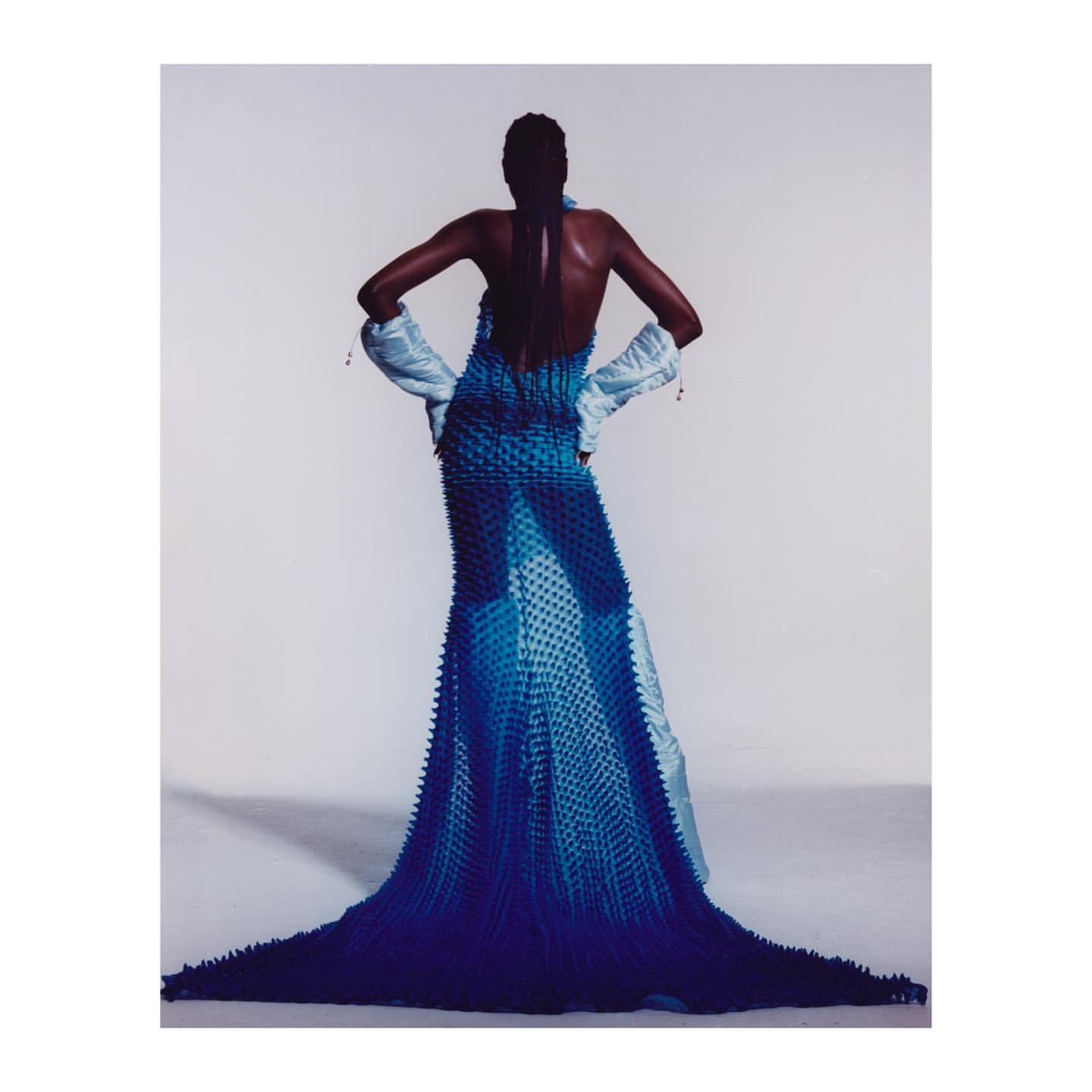 Model in blue textured gown and light blue gloves against a white background, fashion editorial photography.