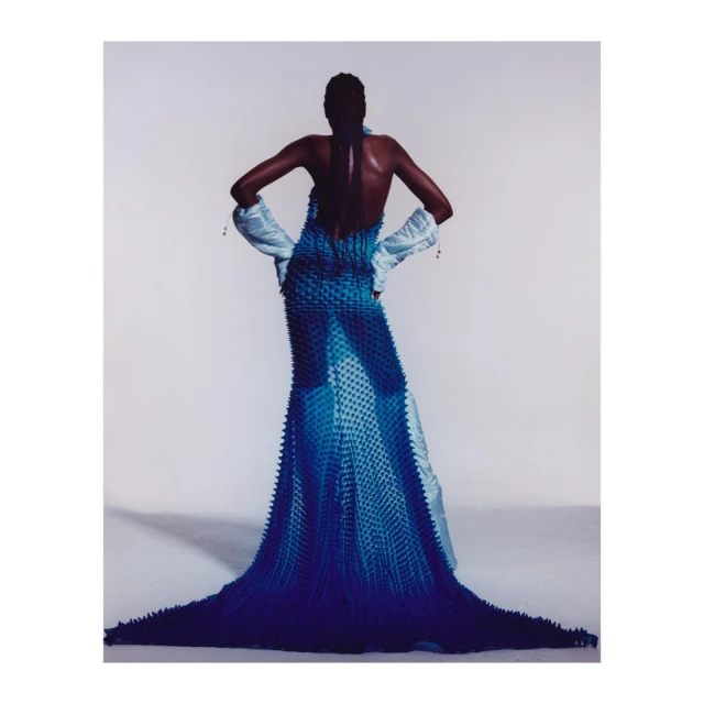 Model in blue textured gown and light blue gloves against a white background, fashion editorial photography.