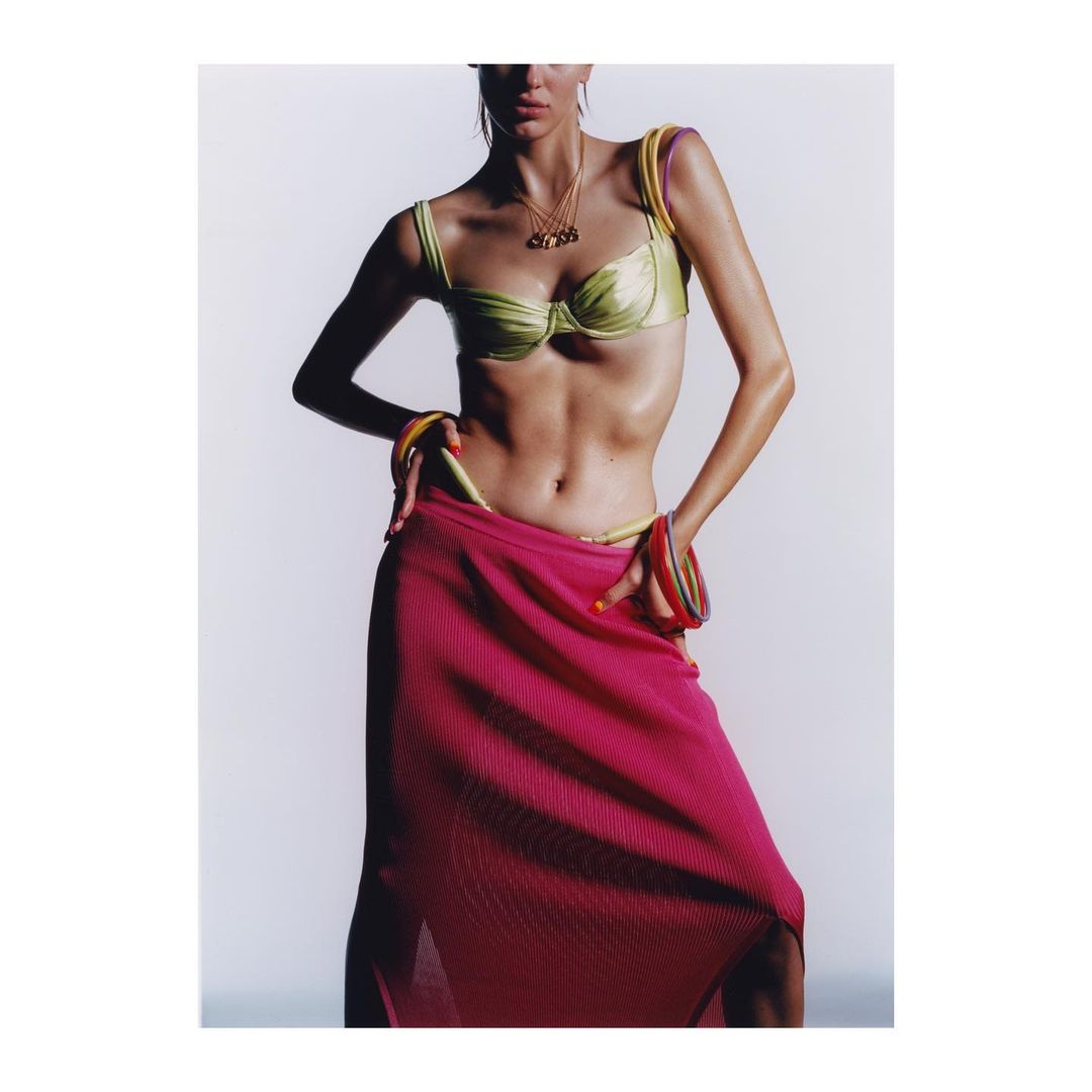 A slender woman poses in a photo studio, showcasing a green bikini top, pink skirt, and colorful bracelets against a white backdrop.