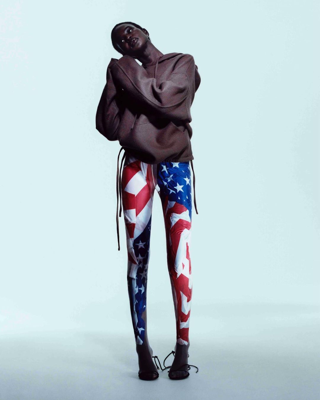 An African American woman poses in a studio wearing an oversized brown hoodie and form-fitting leggings patterned with the American flag.