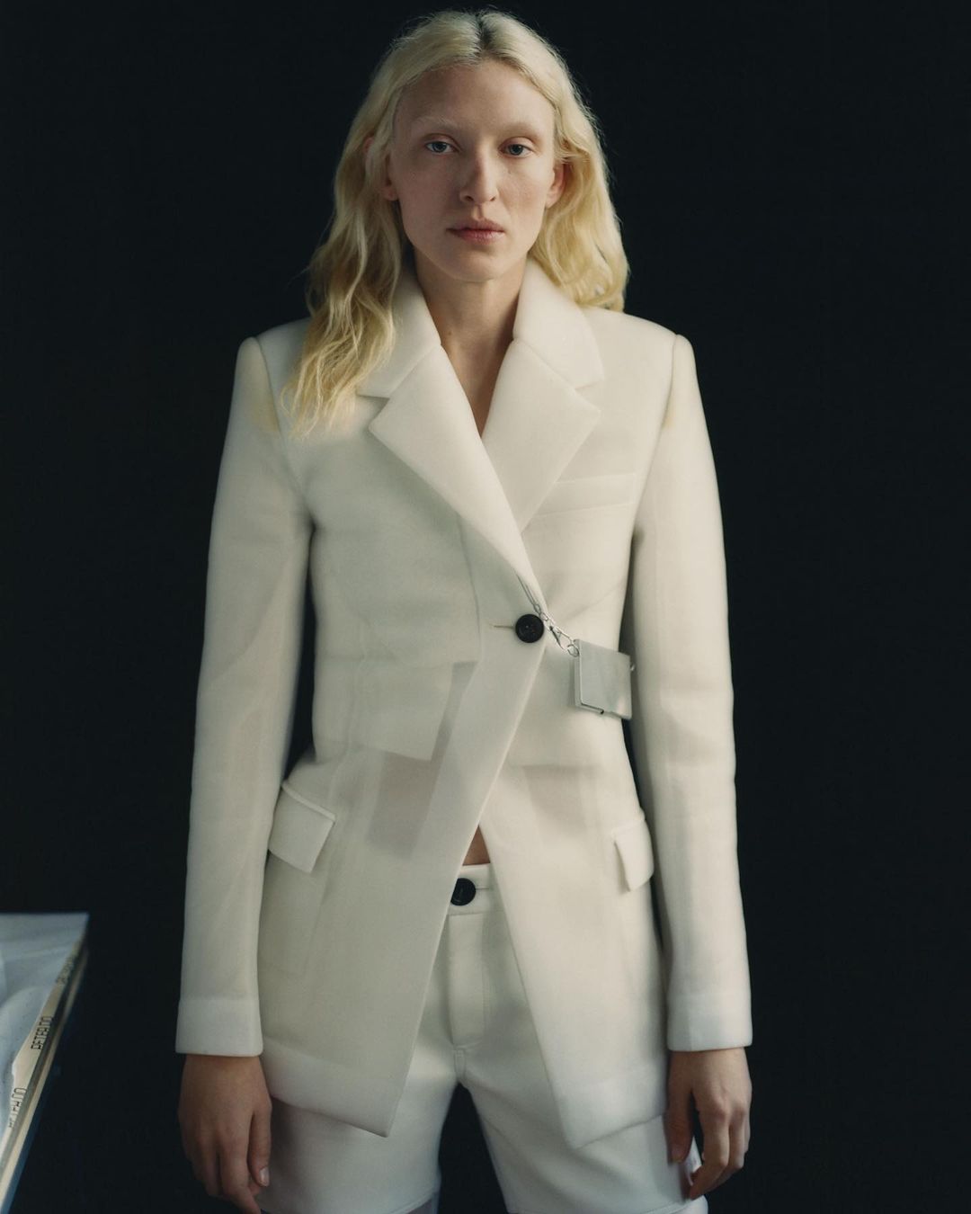Fashion portrait of a woman in a white blazer and shorts against a dark background. Studio lighting highlights the clothing details.