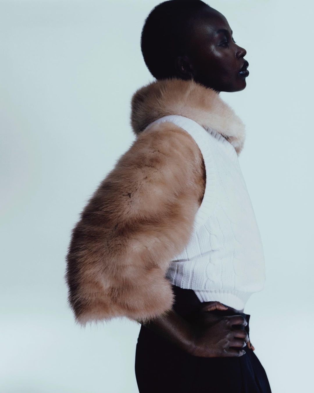 Profile shot of a Black female model in a fur-trimmed vest, hands clasped, against a light backdrop, showcasing fashion and elegance.