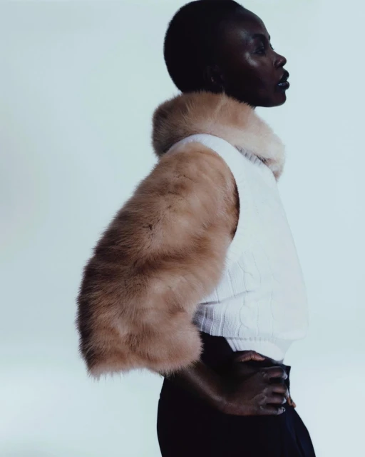 Profile shot of a Black female model in a fur-trimmed vest, hands clasped, against a light backdrop, showcasing fashion and elegance.
