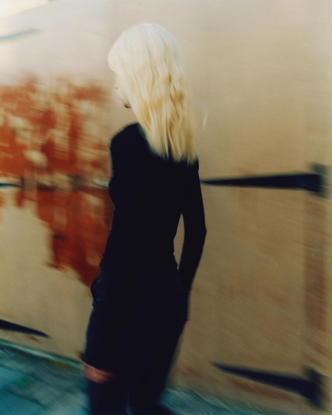 A full body shot of a woman with blonde hair walking towards the left side of the frame in a blurry urban environment.