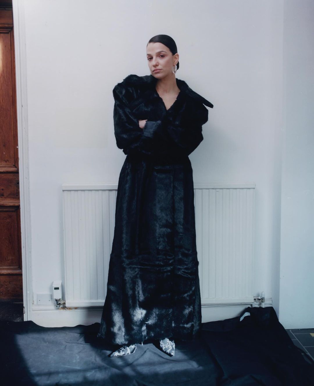 Woman in a black fur coat stands in front of a white wall, posing with her arms crossed in an indoor setting. Fashion photography.
